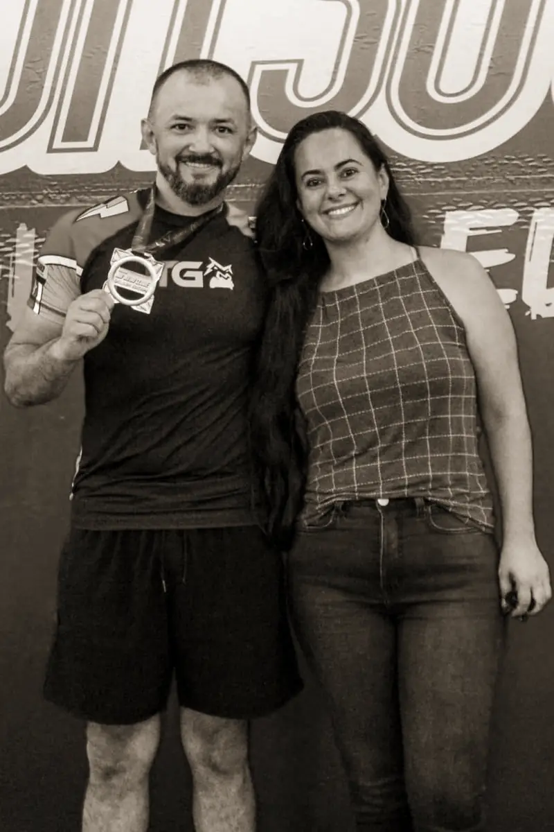 Pécora de rash guard mostrando a medalha ao lado de uma mulher, em frente ao painel da competição