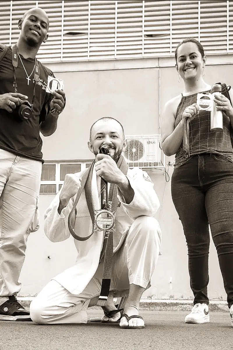 Pécora de kimono, agachado e de punho fechado, com a medalha no peito, entre duas pessoas que seguram medalhas